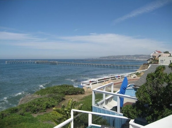 A coastal view with a long pier extending into the sea, a building with a balcony overlooking the ocean, and lush greenery in the foreground.