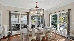 A dining area with a white table set for six, a hanging light fixture, and large windows with patterned curtains. - Short Stays! Family Beach Spot! Dog Friendly! Steps to the BEACH! (Isle of Palms)