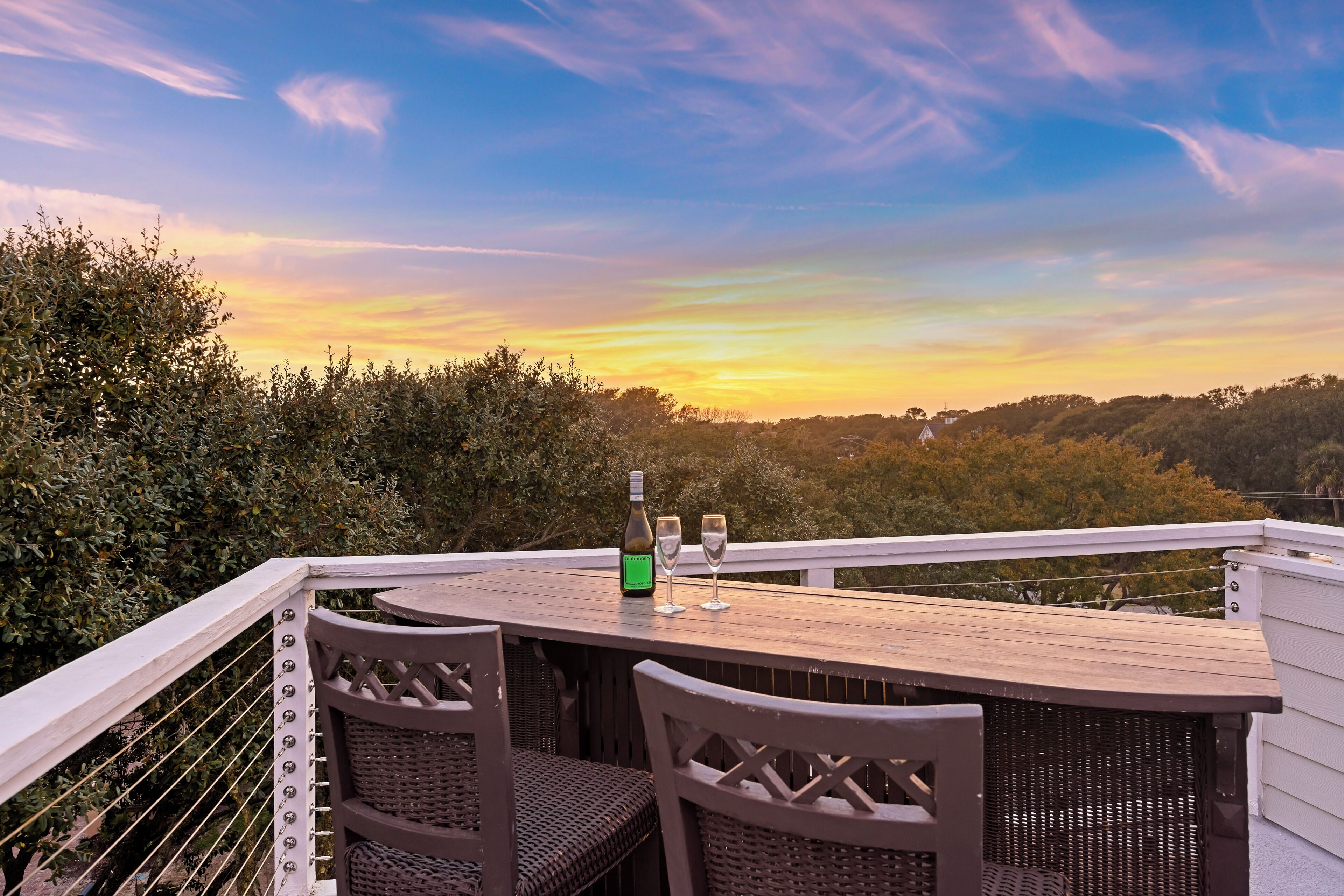 A wooden deck with wicker chairs, a table, and a bottle and glasses, overlooking a forested landscape.