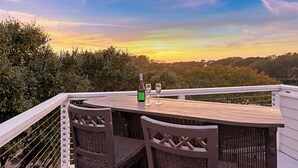 A wooden deck with wicker chairs, a table, and a bottle and glasses, overlooking a forested landscape. - Short Stays! Family Beach Spot! Dog Friendly! Steps to the BEACH! (Isle of Palms)