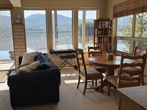 A cozy room with a view of a lake and mountains, a sofa, a dining table, chairs, a keyboard, and a bookshelf.