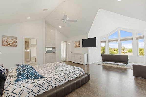 A spacious bedroom with a large bed, a ceiling fan, a flat-screen TV, and a view of the outdoors through large windows.
