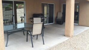 Un patio al aire libre con una mesa y sillas de vidrio, una parrilla y un área de estar cubierta.