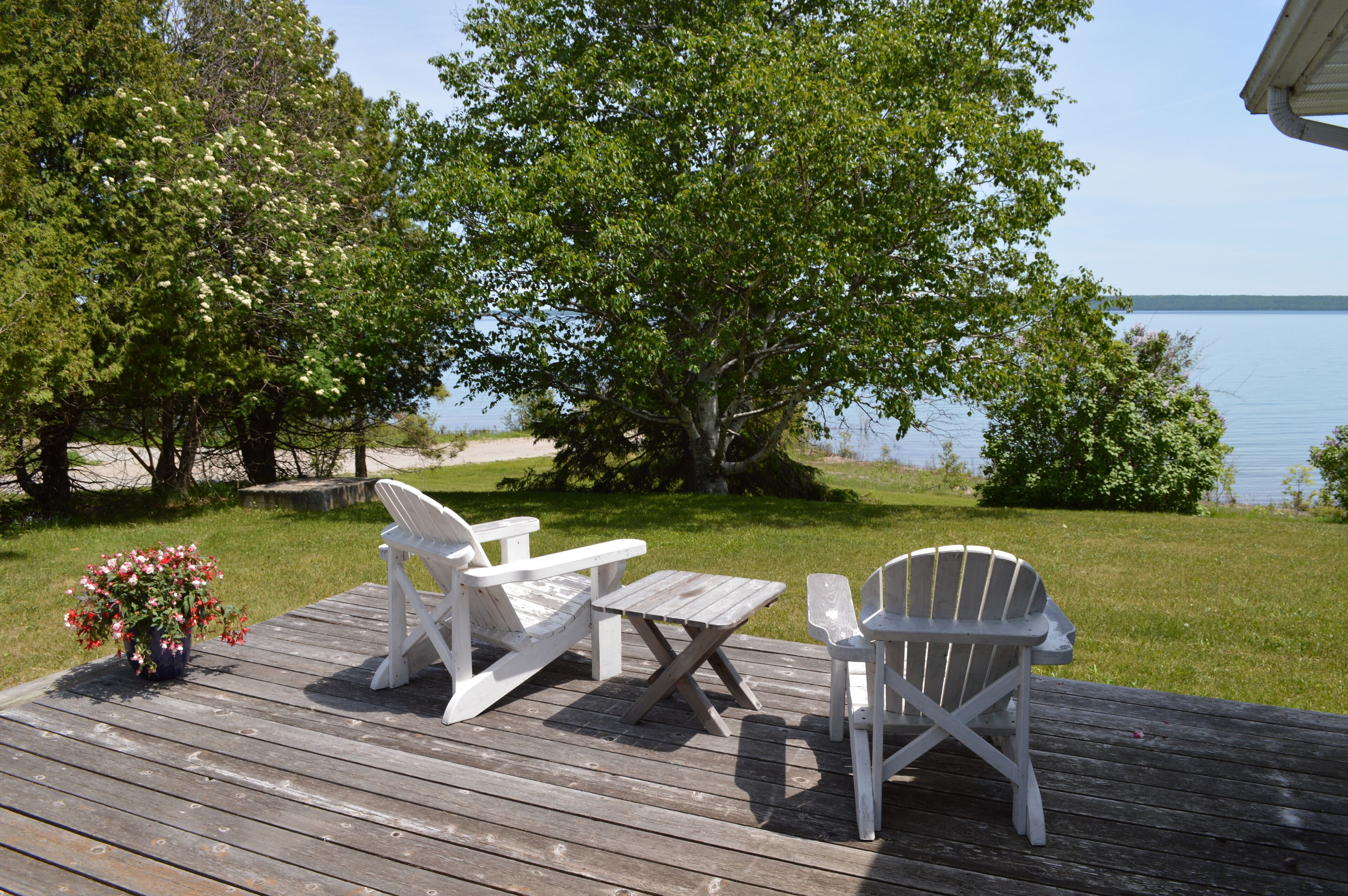 Une terrasse en bois avec deux fauteuils Adirondack et une petite table, donnant sur un lac bordé d’arbres et une zone herbeuse.