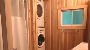 Un baño pequeño de madera con ducha, dos lavadoras apiladas y una ventana pequeña.