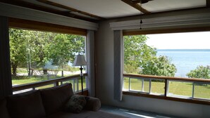 Una habitación con un ventanal grande que da a una masa de agua y follaje. Hay un sofá con cojines y una lámpara cerca de la ventana.