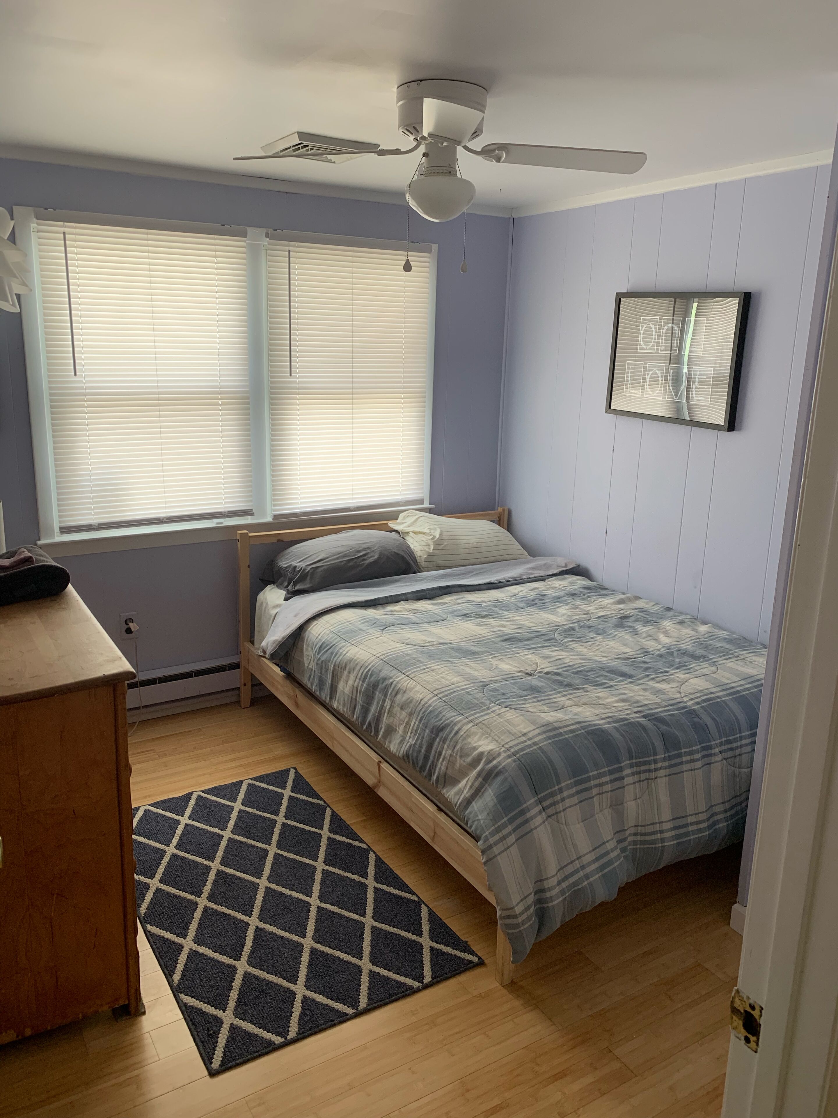 A bedroom with a bed, a ceiling fan, a window with blinds, a wall-mounted picture, and a wooden dresser.