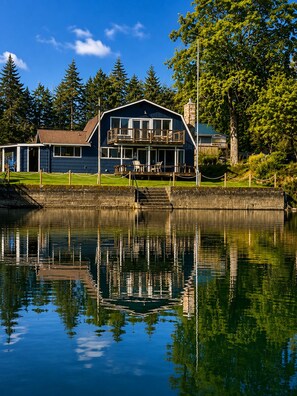 A two-story building with a balcony, situated by a calm body of water, reflecting the structure and surrounding greenery.