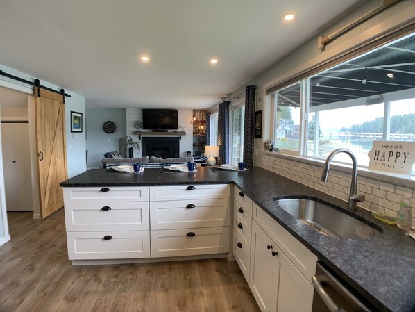 A modern kitchen with a large island, white cabinetry, and a view of a lake through the window. - The Beach Barn - Waterfront Cottage on Case Inlet - Kayaks, Beach, Stunning View (Allyn)