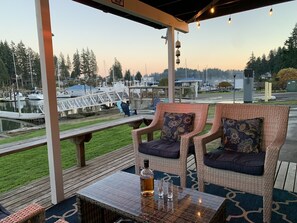 A covered outdoor seating area with wicker chairs and a table, overlooking a marina with boats and docks. - The Beach Barn - Waterfront Cottage on Case Inlet - Kayaks, Beach, Stunning View (Allyn)