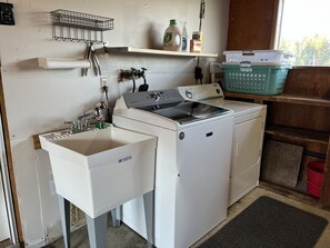 A laundry area with a washing machine, a sink, and a shelf with cleaning supplies. - The Beach Barn - Waterfront Cottage on Case Inlet - Kayaks, Beach, Stunning View (Allyn)