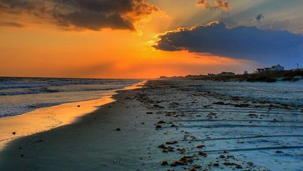 Ein Strand mit Sonnenuntergang, ruhigem Meer und einer fernen Küstenlinie mit Gebäuden.