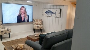 Ein Wohnzimmer mit einem großen Flachbildfernseher, zwei gemusterten Sesseln, einem grauen Sofa, einem Holztisch und einer Wanddekoration mit einem Seestern.