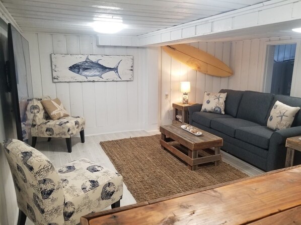 Ein gemütliches Wohnzimmer mit einem grauen Sofa, gemusterten Sesseln, einem Holztisch und einer Surfbrett-Dekoration.