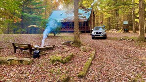 Een blokhut in een bos met een rookspuwend vuurkorf, een bank en een auto die er staat geparkeerd.