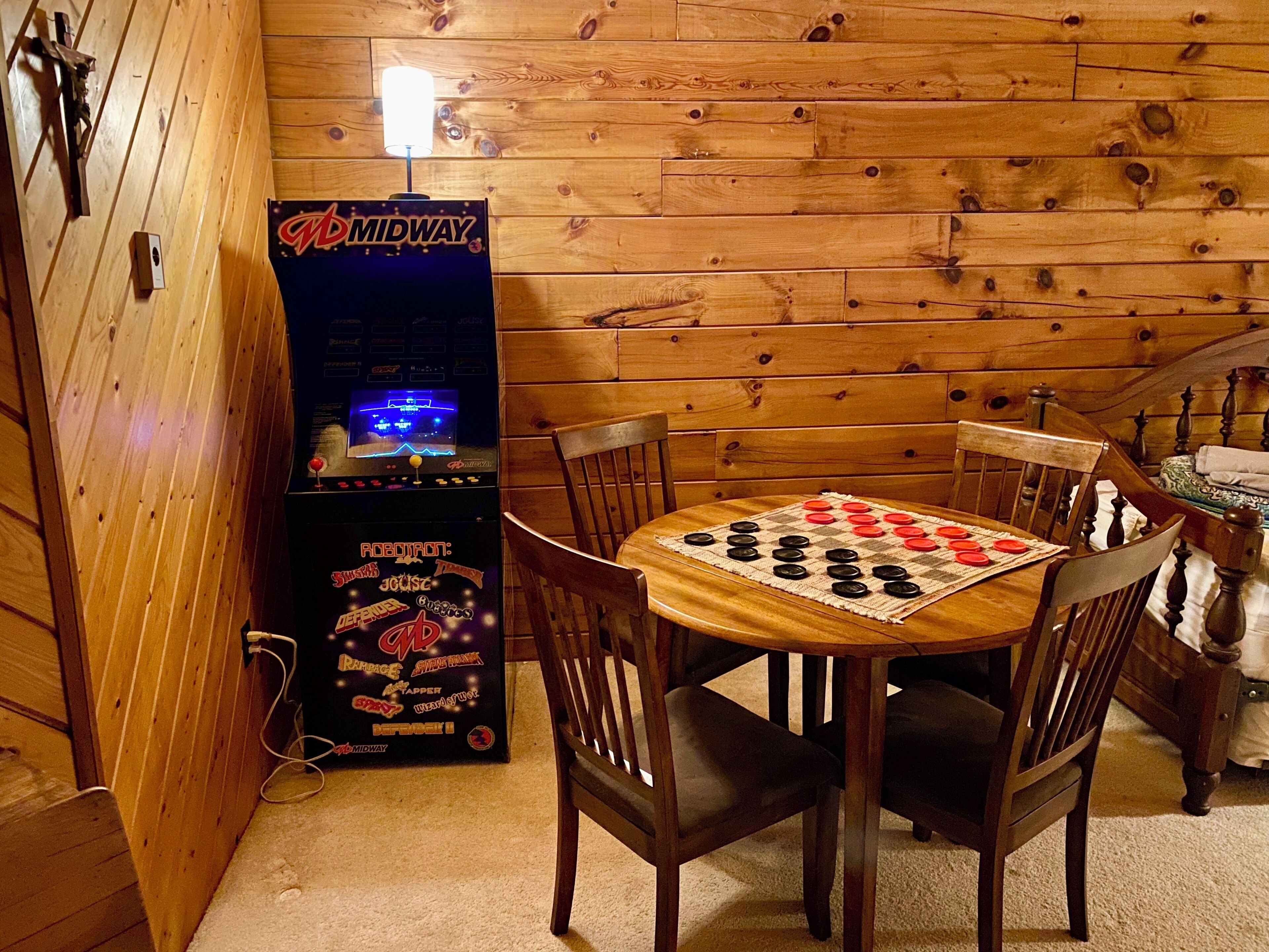 A wooden cabin interior with a Midway arcade game, a checkered board game, and a bed with patterned bedding.