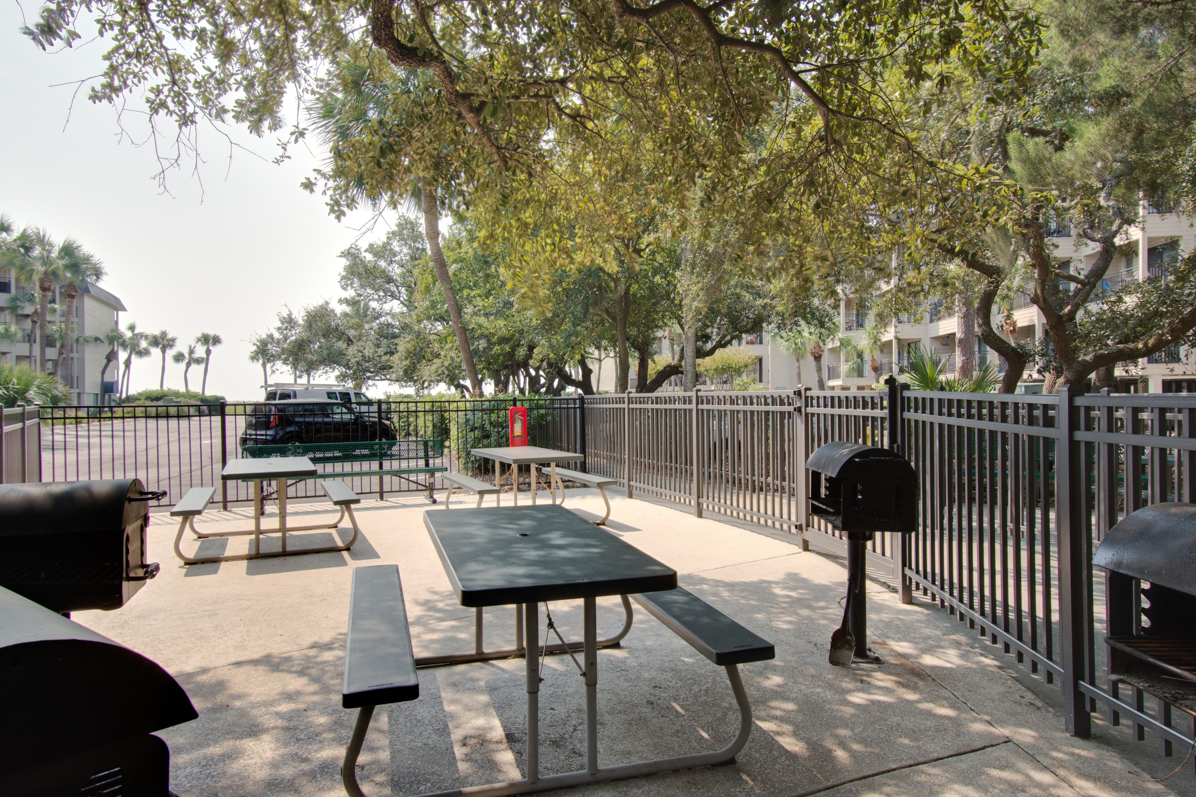 An outdoor patio area with picnic tables and a grill, surrounded by a metal fence and trees.