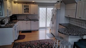 Une cuisine avec des armoires blanches, un dosseret carrelé noir et blanc, une cuisinière à gaz, un évier et un réfrigérateur. Un tapis à motifs recouvre le sol, et un petit tabouret est placé près de l’évier.