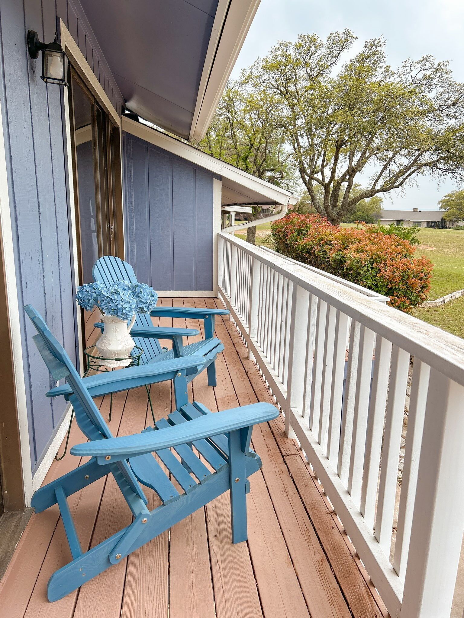 A wooden deck with blue Adirondack chairs and a white vase with flowers.