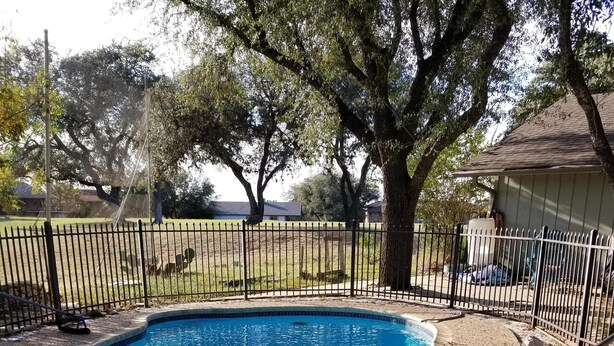 A swimming pool with a surrounding fence, trees, and a building in the background.