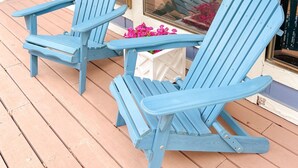 Two blue Adirondack chairs on a wooden deck with a glass door and windows.