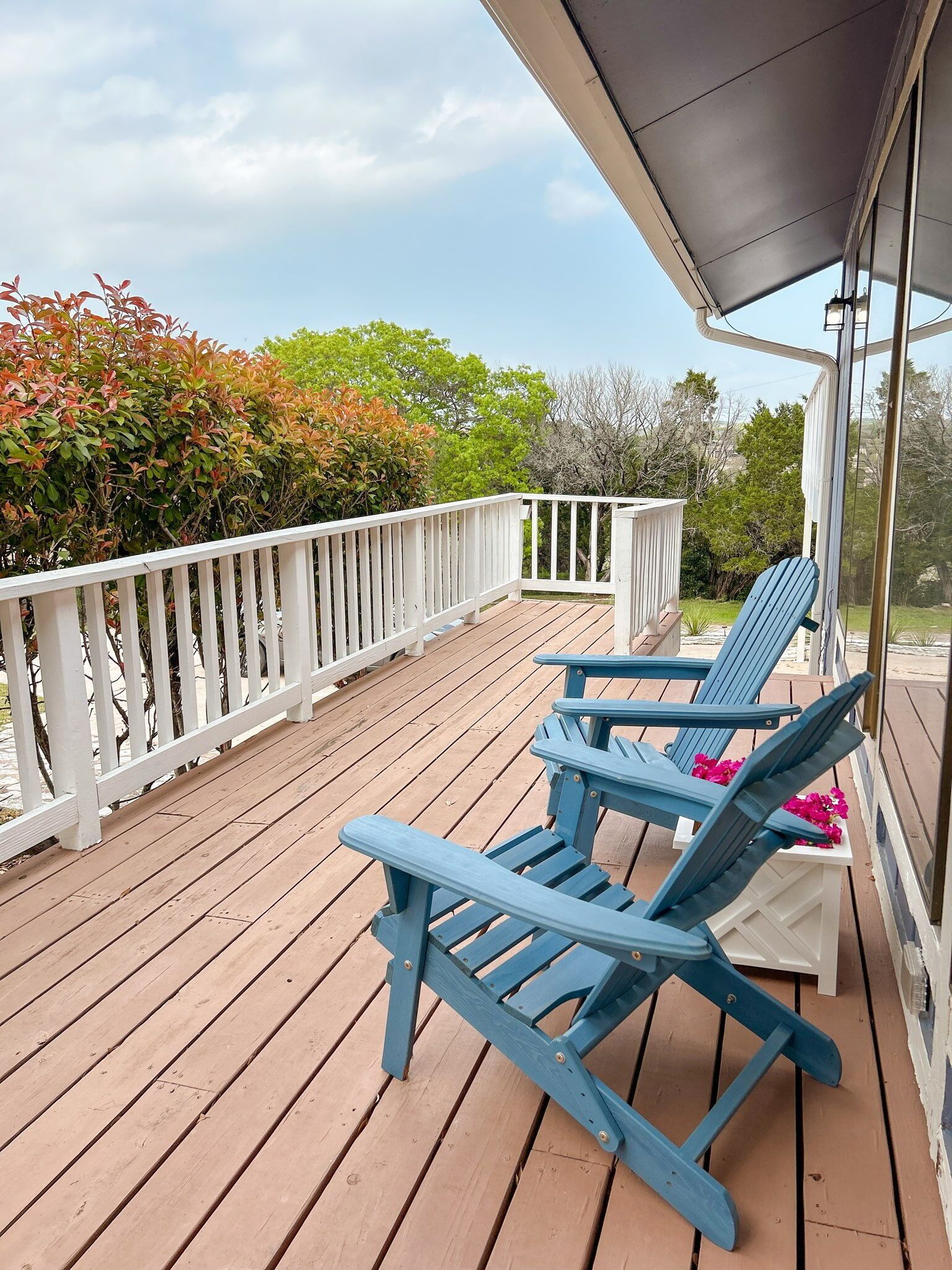 A wooden deck with a white railing, two blue Adirondack chairs, and a small table with pink flowers.