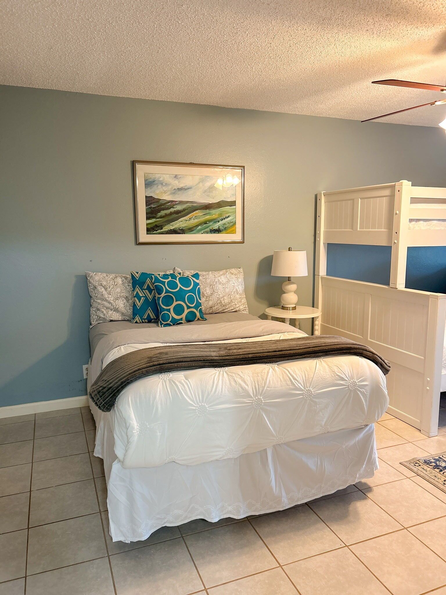 A bedroom with a bed, a nightstand with a lamp, a framed painting on the wall, and a bunk bed in the corner.