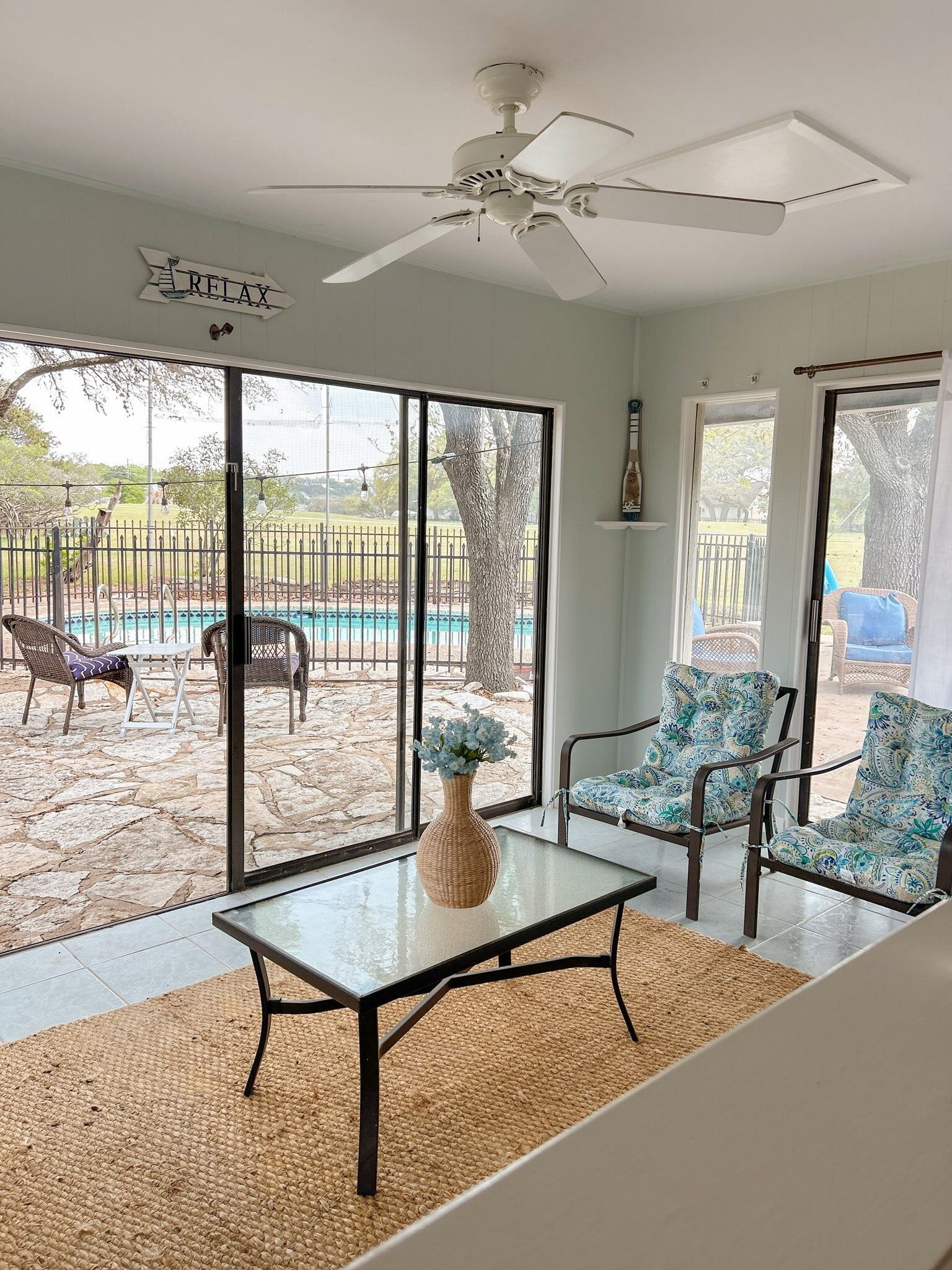 A room with a glass table, two patterned chairs, a ceiling fan, and a view of a swimming pool and outdoor seating area.