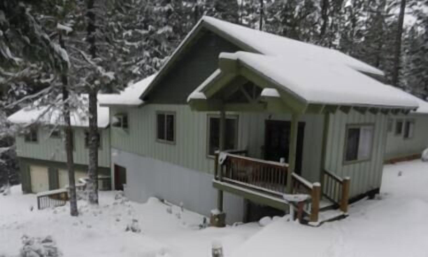 A two-story cabin with a porch and a covered deck, surrounded by snow-covered trees.