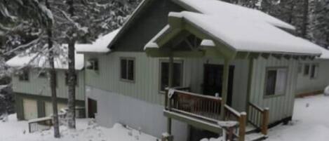 A two-story cabin with a porch and a covered deck, surrounded by snow-covered trees.