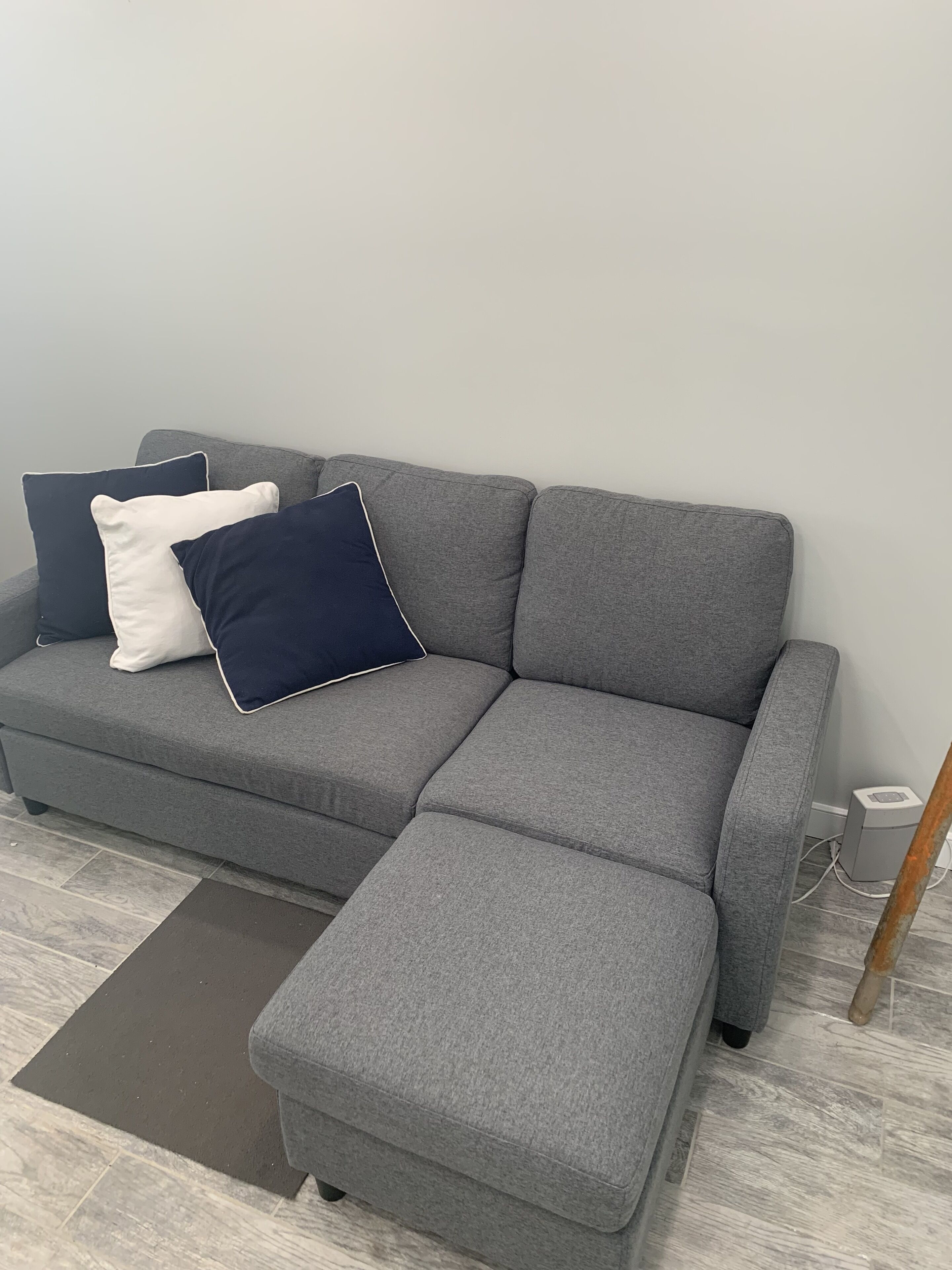 A grey sectional sofa with a matching ottoman, two pillows, and a small speaker unit against a plain wall.