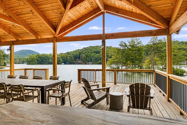 A wooden deck with a table and chairs, overlooking a lake with a forested hill in the background.