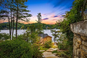 A lakeside cabin with a stone wall, wooden deck, and a staircase leading to the entrance.