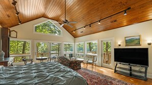 A spacious bedroom with a large bed, a ceiling fan, and a TV. There are multiple windows offering views of the outdoors. - Lake Summit Estate.  Directly On The Water! (Zirconia)