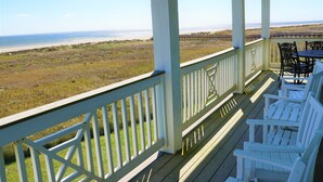 A covered porch with white railings, a table, and four white Adirondack chairs facing a beach.