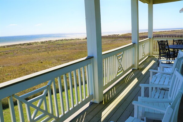 A covered porch with white railings, a table, and four white Adirondack chairs facing a beach.