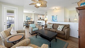 A spacious living room with a ceiling fan, a dining area with chairs, a kitchen in the background, and a view to an outdoor deck.