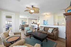 Un vaste salon doté d’un ventilateur de plafond, un coin repas avec des chaises, une cuisine à l’arrière-plan et une vue sur une terrasse extérieure.