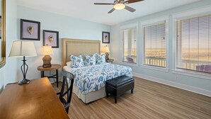 A bedroom with a bed, bedside tables, a desk, a ceiling fan, and large windows with blinds.