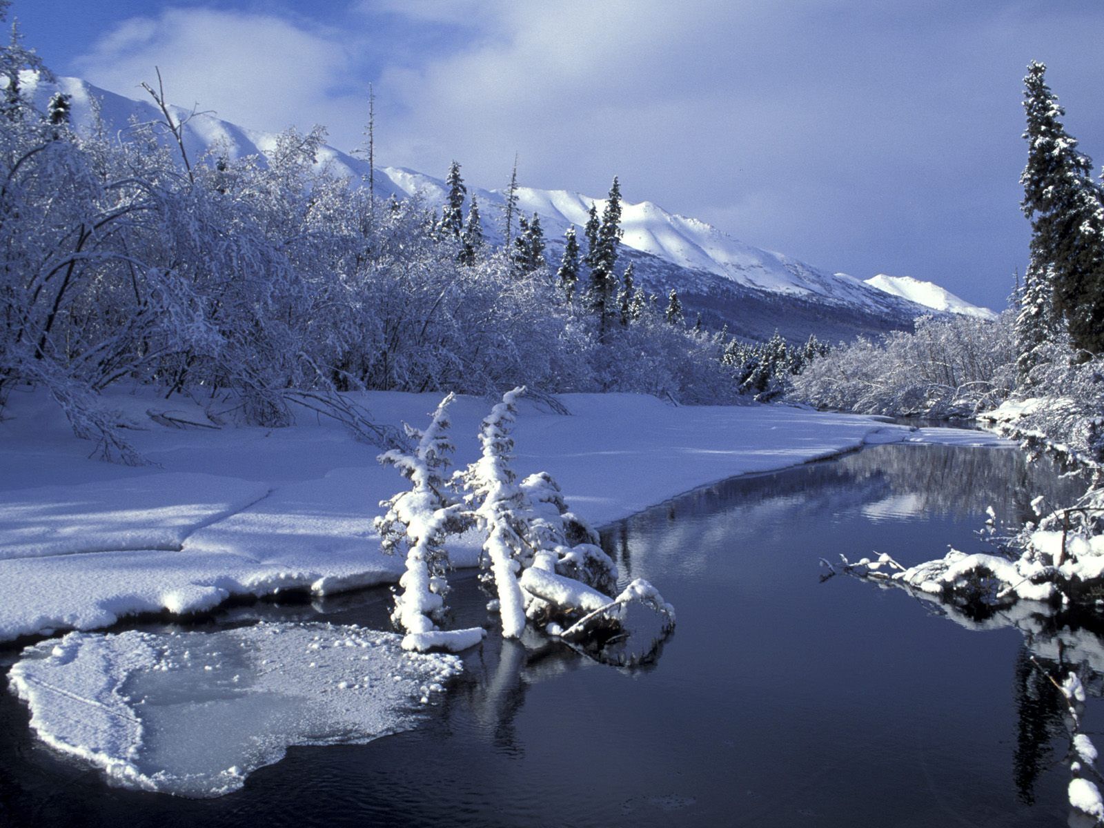 Un paisaje invernal sereno con un río helado, árboles cubiertos de nieve y montañas al fondo.