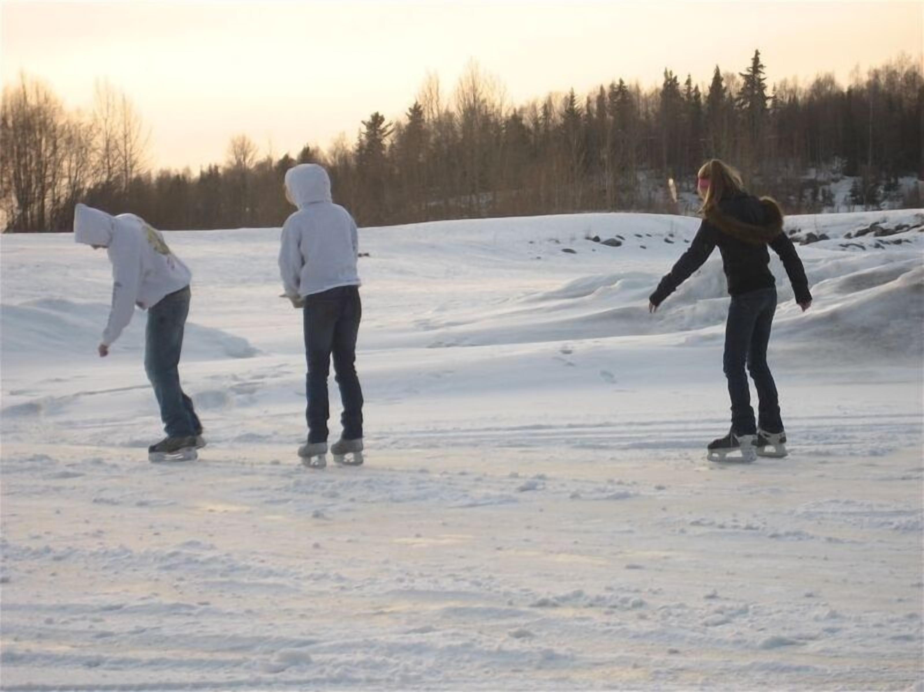 Trois personnes font du patin à glace sur une surface plane et enneigée, avec des arbres en arrière-plan.