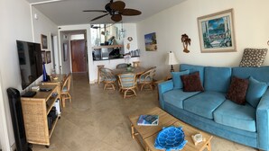A living room with a blue sofa, wooden dining table, and chairs. There is a TV, a ceiling fan, and a lamp.