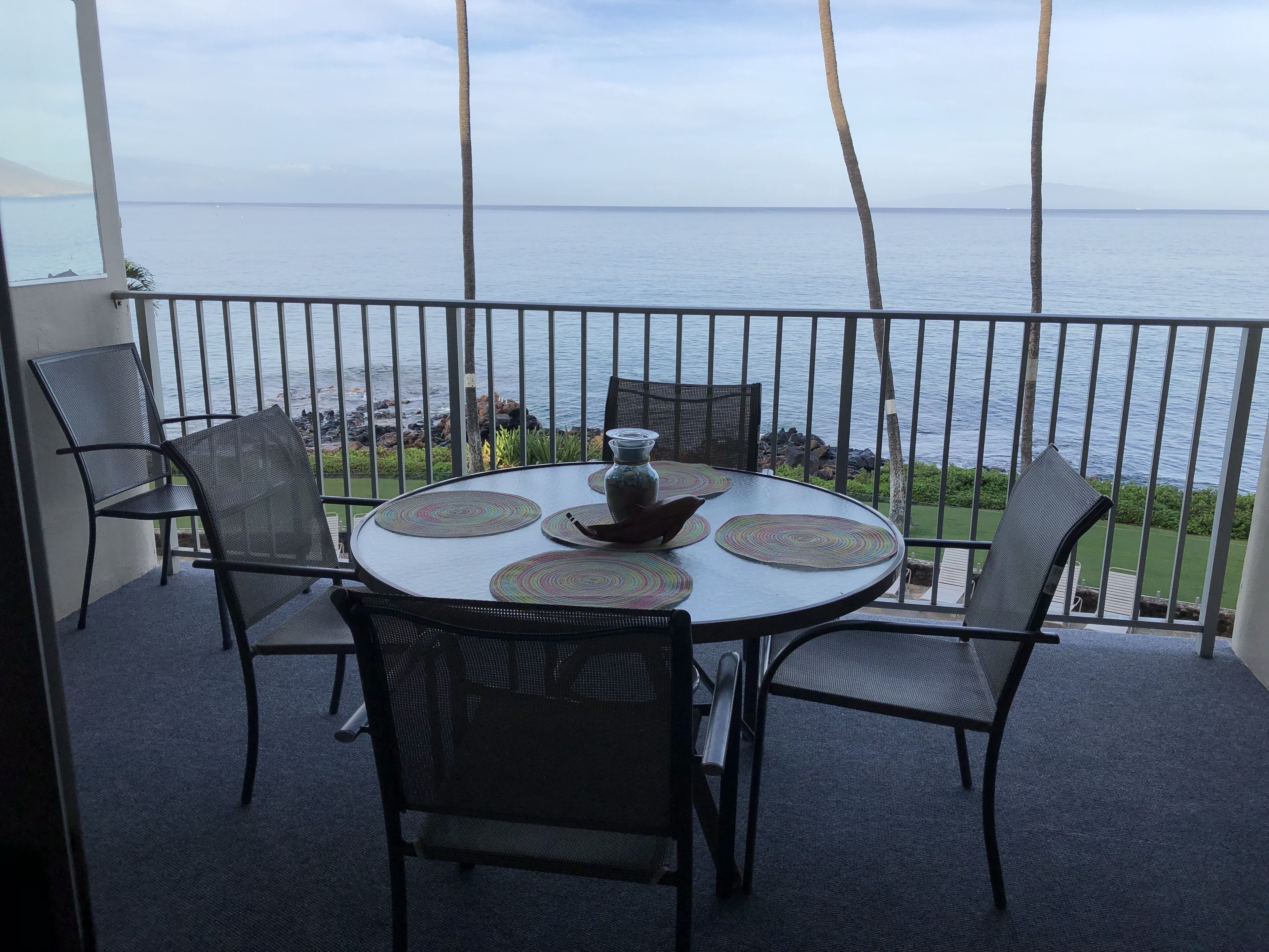 Una mesa redonda con cuatro sillas y un florero, ubicada en un balcón con vistas al mar y a la costa rocosa.
