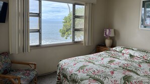 A bedroom with a bed, a chair, a nightstand, a lamp, and a window with a view of the ocean.