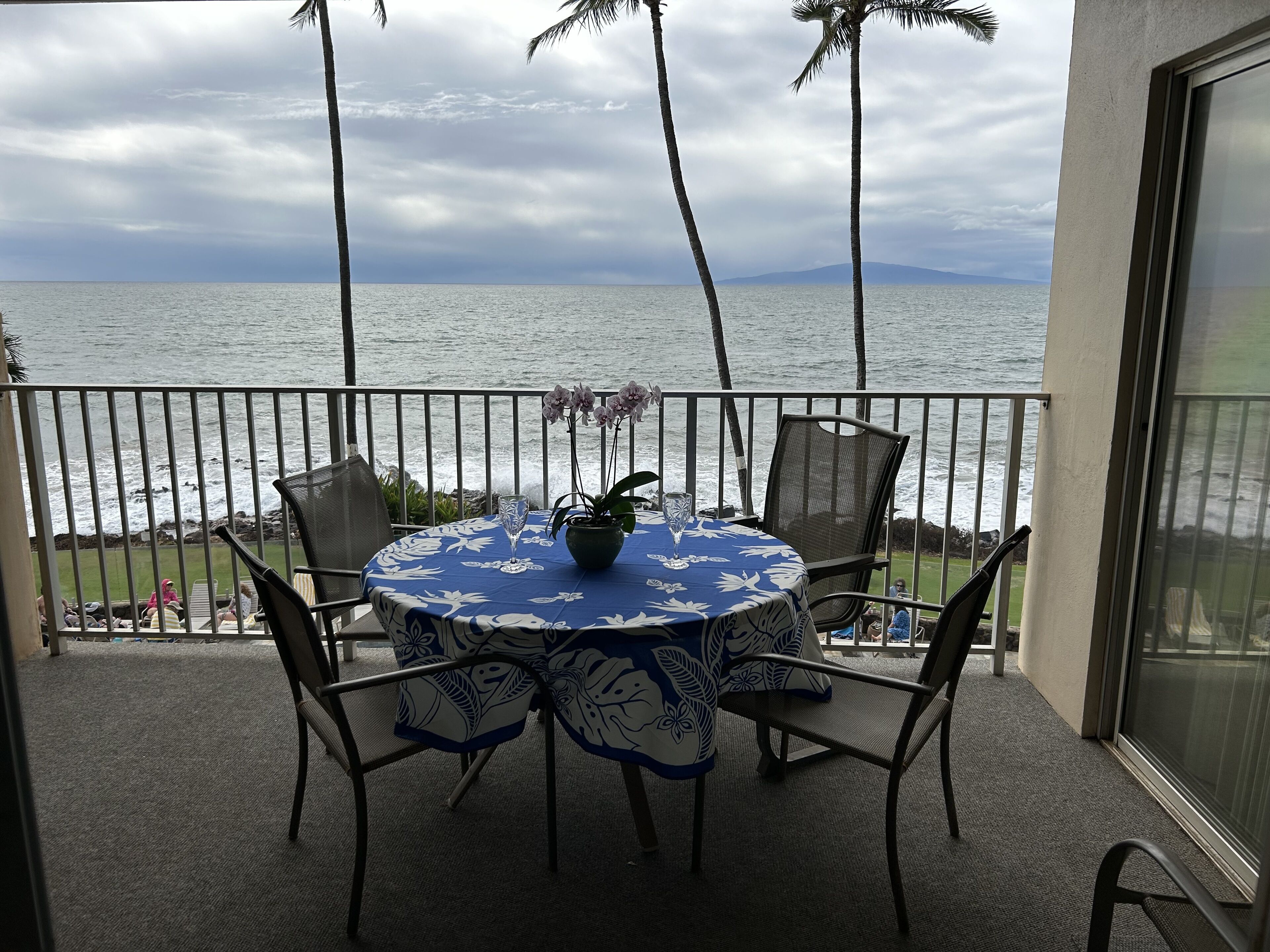 A round table with a floral tablecloth, surrounded by chairs, on a balcony with a view of the ocean and palm trees.