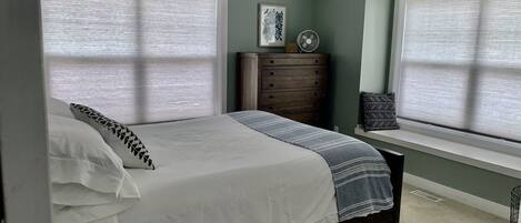 Een slaapkamer met een bed, een dressoir, een raam met jaloezieën en een radiator.