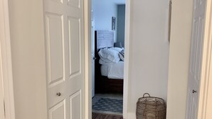 Un pasillo con suelo de madera, una puerta blanca a la izquierda y una cama con cabecera blanca al fondo.