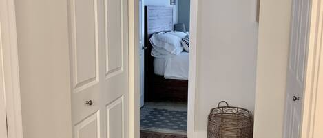 A hallway with a wooden floor, a white door on the left, and a bed with a white headboard in the background.