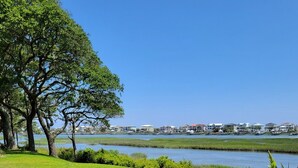 A well-maintained grassy area with a large tree and a small water feature, leading to a view of a coastal town with buildings. - Breathtaking Waterway Views. 1BR. 1Bath. Kitchen. Porch. 2 Person. 5 Star Views (Ocean Isle Beach)