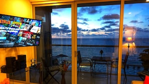 Una sala de estar moderna con un televisor grande que muestra varios servicios de streaming, un balcón con vistas al mar y una zona de estar acogedora con una mesa y sillas.